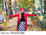 Happy female wearing red beret and coat tossing yellow dry leaves on autumn street and looking at camera. Cheerful woman throwing maple leaves in air. Стоковое фото, агентство Ingram Publishing / Фотобанк Лори