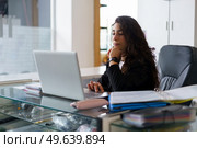 Pensive woman with wavy dark hair touching chin and browsing data on laptop while sitting at desk with sports supplies during work in gym. Thoughtful sports psychologist using netbook during work. Стоковое фото, агентство Ingram Publishing / Фотобанк Лори