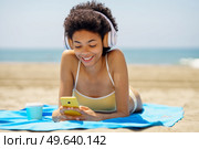Smiling black woman lying on a towel on the sand on the beach using her smartphone to check Social Media. African American girl tanning.. Smiling black woman lying on the beach using her smartphone to check Social Media. Стоковое фото, агентство Ingram Publishing / Фотобанк Лори