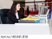 Glad woman with wavy dark hair smiling and typing on netbook keyboard while sitting at desk and working in gym. Sports psychologist using laptop in gym. Стоковое фото, агентство Ingram Publishing / Фотобанк Лори