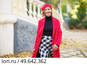 Happy female in red outfit and beret looking at camera while strolling on path near fence on street with trees in city. Smiling woman in stylish outfit walking on street. Стоковое фото, агентство Ingram Publishing / Фотобанк Лори
