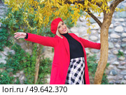 Cheerful young elegant woman with blond hair and red lips in stylish coat and beret holding tree trunk, while standing in autumn park with outstretched arm and smiling. Smiling young female with outstretched arms standing under tree on autumn day. Стоковое фото, агентство Ingram Publishing / Фотобанк Лори