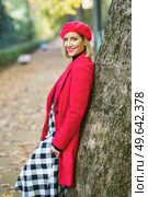Side view of smiling female wearing red outerwear and checkered shirt leaning on tree in autumn park and smiling at camera. Charming woman standing near tree trunk in park. Стоковое фото, агентство Ingram Publishing / Фотобанк Лори