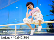 Full body of optimistic African American female looking at camera while sitting on metal fence near glass building in city. Cheerful black woman sitting on railing. Стоковое фото, агентство Ingram Publishing / Фотобанк Лори