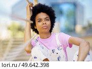 Dreamy young African American female looking away with pensive face while sitting near blurred metal fence on street of city. Wistful black woman sitting near fence. Стоковое фото, агентство Ingram Publishing / Фотобанк Лори