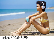Side view of dreamy young African American female tourist with Afro hair, in bikini drinking takeaway coffee and looking away while listening to music in headphones on sandy seashore. Black woman in headphones drinking takeaway beverage while relaxing on beach. Стоковое фото, агентство Ingram Publishing / Фотобанк Лори