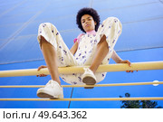 Low angle full body of confident African American female in overall looking at camera while sitting on fence with bent legs. Black woman sitting on railing in city. Стоковое фото, агентство Ingram Publishing / Фотобанк Лори