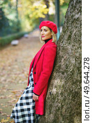 Side view of stylish female wearing red coat, and beret leaning on tree trunk on pathway covered with fallen dry leaves in autumn park and looking away. Serious woman in red outerwear leaning on tree. Стоковое фото, агентство Ingram Publishing / Фотобанк Лори