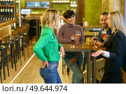 Blond female with glass of wine telling story to diverse male and female friends while gathering around table in bar together. Blond woman speaking with friends in pub. Стоковое фото, агентство Ingram Publishing / Фотобанк Лори