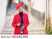 Confident young positive woman, with blond hair in fashionable red coat and beret smiling and looking away while standing on stone stairs with photo camera during trip in old town. Cheerful female traveler with camera smiling on narrow street during vacation. Стоковое фото, агентство Ingram Publishing / Фотобанк Лори