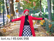 Delighted female wearing red outerwear throwing dry autumn leaves while standing on street in fall season and looking at camera. Happy woman throwing autumn leaves in park. Стоковое фото, агентство Ingram Publishing / Фотобанк Лори