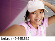 Chinese girl taking a photo or video selfie with her smartphone, to post on social media. Young Asian woman wearing modern clothes and cap.. Asian girl taking a photo or video selfie with her smartphone, to post on social media. Стоковое фото, агентство Ingram Publishing / Фотобанк Лори