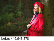 Side view of stylish female wearing red beret and jacket leaning on tree trunk and closing eyes from sunlight in park. Gentle female wearing red beret and jacket standing near tree. Стоковое фото, агентство Ingram Publishing / Фотобанк Лори