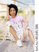 Wistful African American female in casual wear looking away with thoughtful face while sitting near metal fence on street of city. Dreamy black woman sitting near fence. Стоковое фото, агентство Ingram Publishing / Фотобанк Лори
