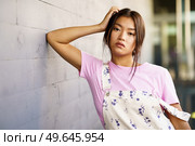Attractive Chinese girl looking at camera with serious expression leaning against the wall of a modern office building. Female wearing casual clothes.. Attractive Chinese girl looking at camera with serious expression leaning against an urban wall. Стоковое фото, агентство Ingram Publishing / Фотобанк Лори