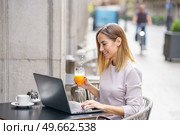 Side view of positive adult self employed lady with blond hair in casual clothes, drinking glass of fresh juice and smiling while working remotely on laptop in street cafe. Happy female freelancer working on netbook in outdoor cafeteria. Стоковое фото, агентство Ingram Publishing / Фотобанк Лори