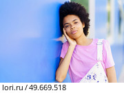 Calm young African American female in casual wear looking at camera and touching cheek while standing near blue building in city. Charming black woman standing near house on street. Стоковое фото, агентство Ingram Publishing / Фотобанк Лори