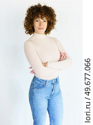 Self assured female model with dark curly hair in beige turtleneck and jeans crossing hands while standing against white background. Confident curly haired woman with crossed arms. Стоковое фото, агентство Ingram Publishing / Фотобанк Лори