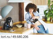 Curly haired female in unbuttoned shirt and black bra pouring hot tea into mug while leaning on electric stove. Woman pouring tea from french press into cup in modern kitchen. Стоковое фото, агентство Ingram Publishing / Фотобанк Лори