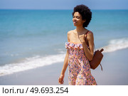 Black woman in colorful dress smiling while exploring sandy beach near rippling ocean and looking away. Young ethnic female spending summer vacation on seashore. Стоковое фото, агентство Ingram Publishing / Фотобанк Лори