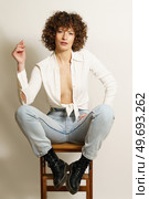 Full length of flirty female model with curly hair, in stylish wear resting on wooden stool playing with curl and looking at camera against white background. Self confident woman chilling on ladder. Стоковое фото, агентство Ingram Publishing / Фотобанк Лори