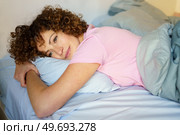 Positive young woman with curly hair sleeping on comfortable bed looking away dreamily in bedroom at home. Content woman resting in bedroom at home. Стоковое фото, агентство Ingram Publishing / Фотобанк Лори