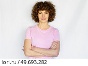 Curly haired woman in casual clothes standing with crossed hands and looking at camera while smiling in light studio. Adult woman with curly hair and crossed arms against white background. Стоковое фото, агентство Ingram Publishing / Фотобанк Лори