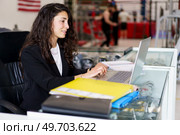 Glad female with dark wavy hair smiling and using laptop while sitting at desk during psychology session in gym. Cheerful sports psychologist using netbook in gym. Стоковое фото, агентство Ingram Publishing / Фотобанк Лори
