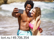 Happy young multiethnic couple in swimwear hugging while taking selfie on mobile phone for social media during honeymoon. Smiling diverse couple taking selfie near sea. Стоковое фото, агентство Ingram Publishing / Фотобанк Лори