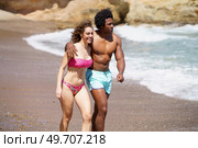 Cheerful multiracial young couple in swimwear smiling and hugging each other while walking together on sandy coast during vacation. Happy diverse man and woman embracing each other while strolling along beach. Стоковое фото, агентство Ingram Publishing / Фотобанк Лори