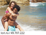Cheerful young African American man with Afro hair giving piggyback ride to happy girlfriend on beach during honeymoon. Black man carrying girlfriend near sea. Стоковое фото, агентство Ingram Publishing / Фотобанк Лори