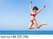 Smiling carefree young female with curly red hair in pink bikini, raising arms and jumping with excitement in air while looking down in daylight against blurred sea and blue sky. Happy woman in bikini jumping with raised arms. Стоковое фото, агентство Ingram Publishing / Фотобанк Лори