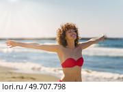 Cheerful young female redhead in pink bikini with eyes closed chin up and smiling with outstretched arms, in sunlight while standing on sandy beach against blue seawater and sky. Happy woman in bikini standing on beach. Стоковое фото, агентство Ingram Publishing / Фотобанк Лори