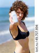 Smiling young female in bikini standing on sandy beach near waving sea and showing white bottle of sunblock cream. Smiling woman showing blank bottle of sunscreen near ocean. Стоковое фото, агентство Ingram Publishing / Фотобанк Лори