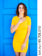 Dreamy young female in yellow dress with curly brown hair keeping hand on chest and looking up against blue wall on street. Charming woman touching chest and looking up. Стоковое фото, агентство Ingram Publishing / Фотобанк Лори