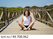 Young curly haired female in casual clothes sitting on wooden walkway and meditating, with closed eyes while doing lotus pose against blurred green hill under blue sky. Relaxed young female meditating on wooden path. Стоковое фото, агентство Ingram Publishing / Фотобанк Лори