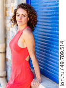 Beautiful young female in red sundress looking away while leaning on stone wall with hand against blue shutters in daylight. Attractive woman in red dress looking at camera. Стоковое фото, агентство Ingram Publishing / Фотобанк Лори