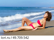 Full length of calm curly woman in bikini and with closed eyes lying on sand of coast of suntanning in sunshine. Sensual lady in bikini sunbathing on coast. Стоковое фото, агентство Ingram Publishing / Фотобанк Лори