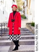 Full length of happy adult female tourist in elegant coat and beret with photo camera on neck smiling and looking away while standing on stone stair in old town. Stylish woman in red coat standing on city street in sunlight. Стоковое фото, агентство Ingram Publishing / Фотобанк Лори