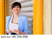 Portrait of positive female in elegant suit smiling and looking away while standing with tablet against silver rolling door with yellow pillars in city. Smiling woman with tablet by rolling door on street. Стоковое фото, агентство Ingram Publishing / Фотобанк Лори