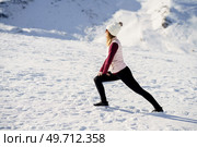 Side view of young female in warm clothes and cap, standing and stretching leg in warrior pose with hands on knee over snowy slope in winter. Woman stretching legs in snowy terrain. Стоковое фото, агентство Ingram Publishing / Фотобанк Лори