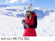 Smiling young female traveler in warm clothes beanie wool cap polarized sunglasses, looking down and opening thermos water bottle while standing on snowy mountain slope in sunny winter day. Happy woman standing with water bottle in sunlight on snowy terrain. Стоковое фото, агентство Ingram Publishing / Фотобанк Лори