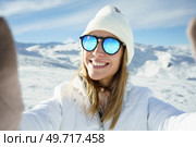 Smiling young female blonde in warm clothes beanie wool cap and polarized sunglasses, looking at camera and taking selfie while standing on snowy mountain slope on sunny day in winter. Cheerful woman in sunglasses taking selfie in snowy place. Стоковое фото, агентство Ingram Publishing / Фотобанк Лори