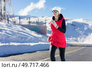 Smiling young female in red vest beanie cap polarized sunglasses gloves looking away, while jogging on asphalt road near snow against snowy mountain and blue sky in daylight. Happy young female in warm clothes and cap running on road. Стоковое фото, агентство Ingram Publishing / Фотобанк Лори
