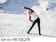 Full body of young female in cap polarized sunglasses, looking away and stretching arm sideways while standing with legs apart and hand on thigh on snowy mountain in daylight. Young woman standing and doing exercise on snow in highlands. Стоковое фото, агентство Ingram Publishing / Фотобанк Лори