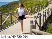 Full body of smiling young female with hands on waist eyes closed standing with stretched leg on bridge fence, while hearing music from earbuds in sunlight against blurred nature. Happy young woman warming up with stretching leg on bridge. Стоковое фото, агентство Ingram Publishing / Фотобанк Лори