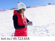 Side view of young female in warm clothes, beanie cap polarized sunglasses looking down at wristwatch while standing on snowy mountain slope in daylight against blurred landscape and blue sky. Happy young woman standing and checking time on wristwatch. Стоковое фото, агентство Ingram Publishing / Фотобанк Лори