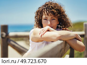 Calm young female with curly hair in white outfit leaning against fence and looking at camera while relaxing alone near blurred background of sea. Young woman leaning on wooden handrail at seaside. Стоковое фото, агентство Ingram Publishing / Фотобанк Лори
