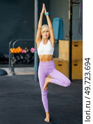 Full body of barefoot female athlete in leggings and top doing Vrikshasana, with arms lifted above head and palms together in prayer position during yoga training. Fit blonde female doing Tree with Arms Up pose in gym. Стоковое фото, агентство Ingram Publishing / Фотобанк Лори