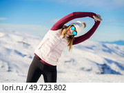 Positive young female in sportswear and sunglasses stretching arms sideway in yoga pose and looking at camera while standing on background of snowy mountains. Cheerful woman stretching arms sideway on snowy terrain. Стоковое фото, агентство Ingram Publishing / Фотобанк Лори